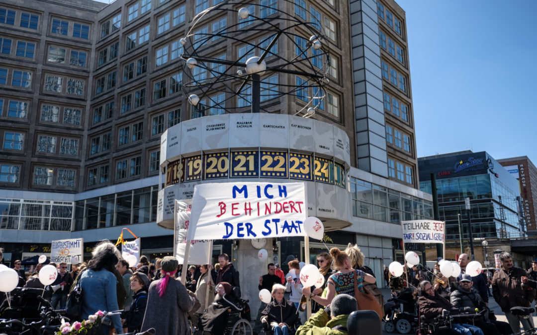 Vor der Weltzeituhr in Berlin demonstrieren mehrere Menschen. Auf einem Transparent steht: Mich behindert der Staat.