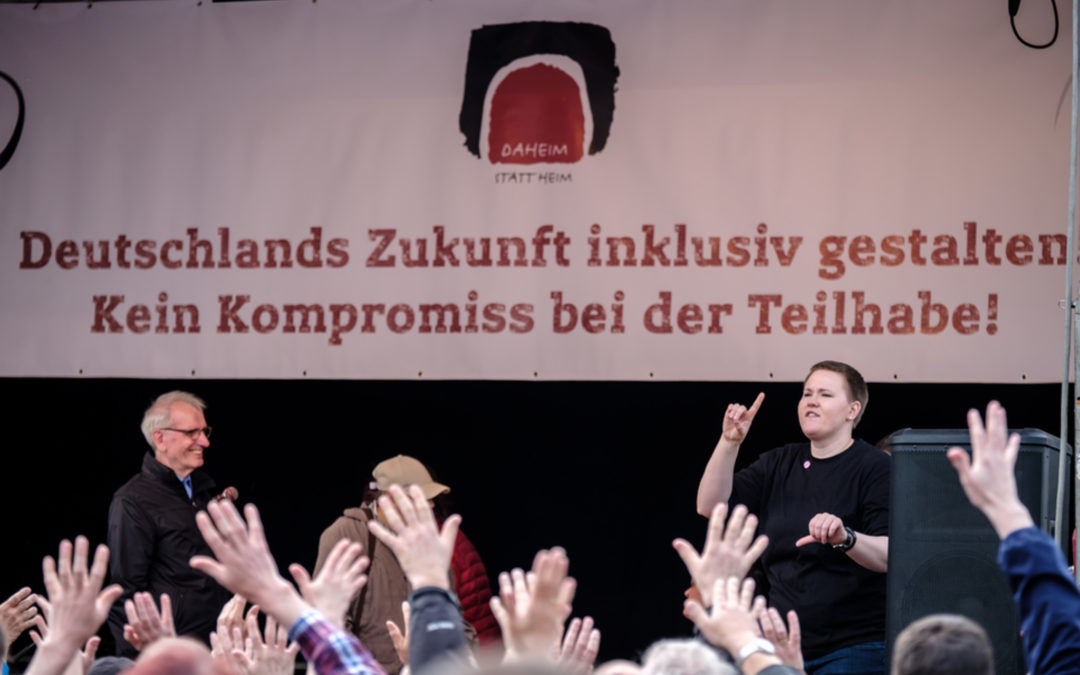 Foto von einer Kundgebung. Auf der Bühne hängt ein Plakat mit den Worten: Deutschlands Zukunft inklusiv gestalten. Kein Kompromiss bei der Teilhabe!