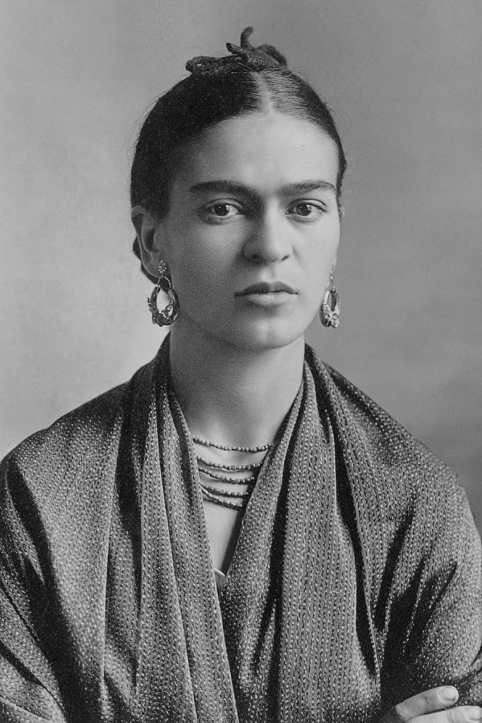 Schwarz-weiß Foto von Frida Kahlo, einer jungen Frau mit zu einem Dutt zusammengebundenen Haaren. Sie trägte große Ohringe und eine Kette und schaut in die Kamera.