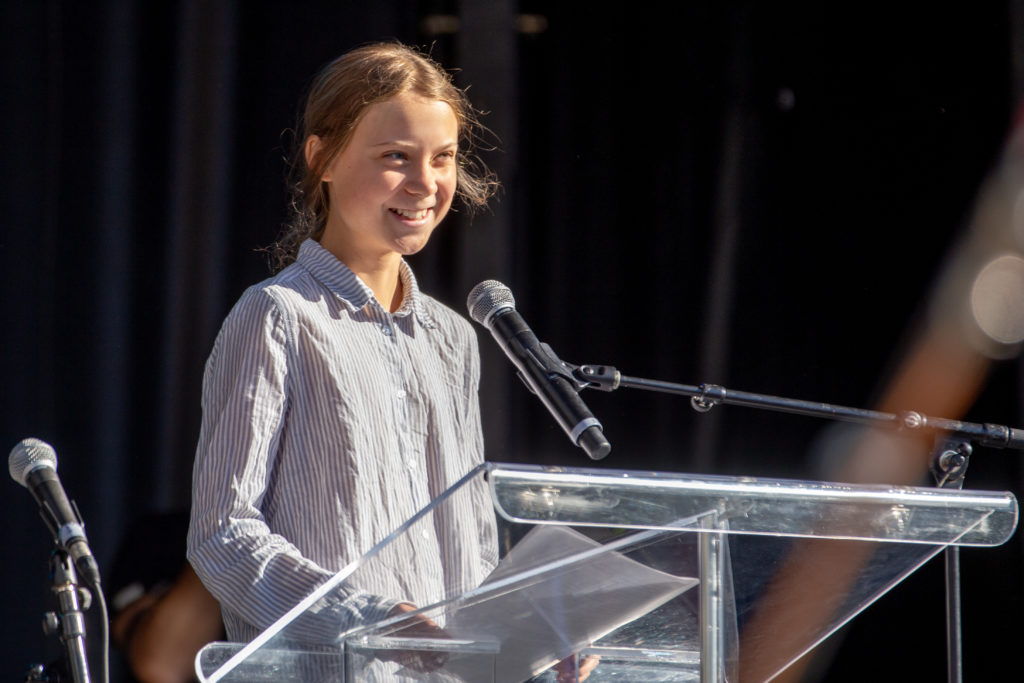 Greta Thunberg steht an einem Rednerinpult und lächelt