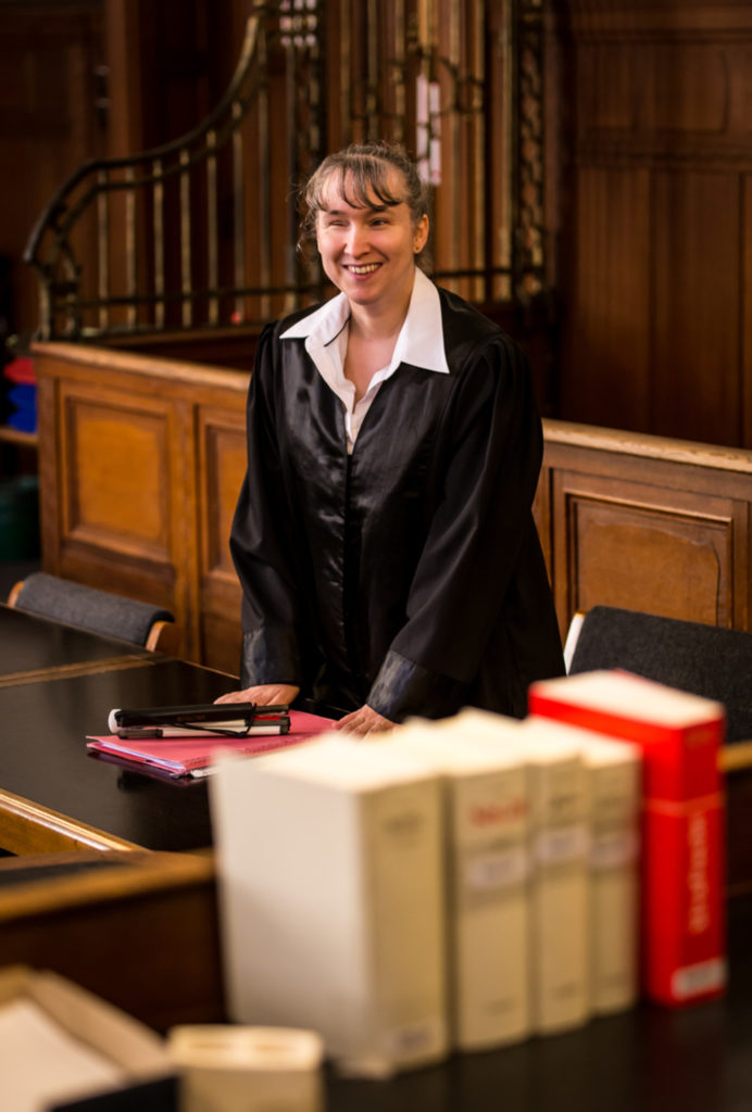 Foto von Pamela Pabst. Sie steht im Gerichtssaal hinter einem Tisch, auf dem viele Bücher und Akten liegen. Sie trägt eine schwarze Anwaltsrobe und hat lange graue Haare.