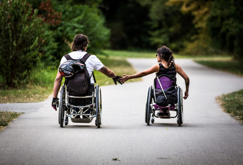 Ein Rollstuhlfahrer und eine Rollstuhlfahrerin von hinten, sie halten Händchen.