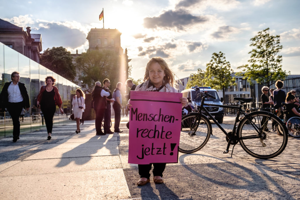 Eine kleinwüchsige Frau steht auf dem Bürgersteig und hält ein pinkes Schild hoch. Darauf steht: Menschenrechte jetzt!