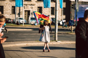 Ein junges Mädchen in einem weißen Sommerkleid ist von hinten zu sehen. Sie szeht auf der Straße und schwenkt eine bunte Fahne.