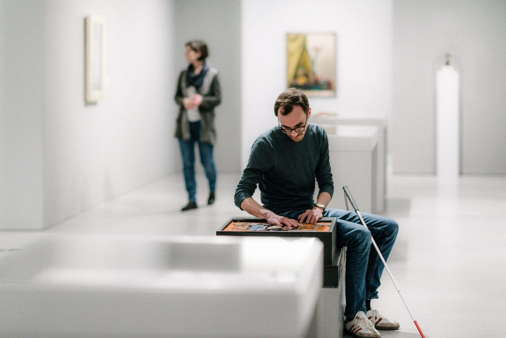 Andreas sitzt in einem weißen Ausstellungsraum und tastet mit seinen Händen einen kunstgegenstand ab.