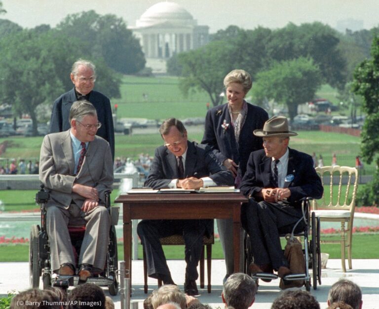 Im Garten des weißen Hauses in Washington sitzt George H.W. Bush an einem Tisch und unterschreibt den Americans with Disabilities Act (ADA). Neben ihm sitzen zwei Personen im Rollstuhl. dahinter stehen zwei weitere Personen.