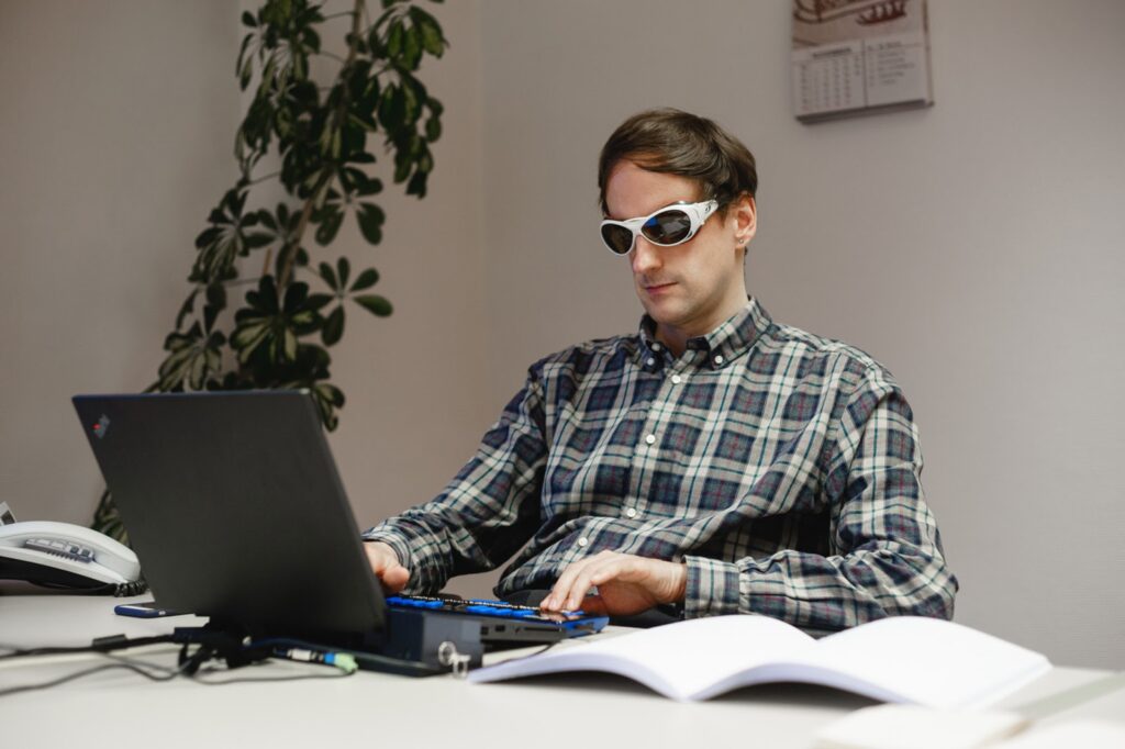 Ein weißer Mann mit Sonnenbrille sitzt am Laptop und bedient eine Braillezeile