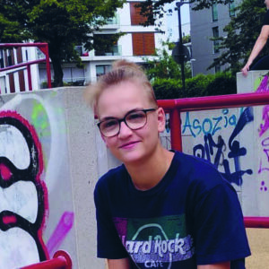 Paulina hat kurze blonde Haare, trägt ein schwarzes T-Shirt und sitzt vor einer Mauer mit Graffitti