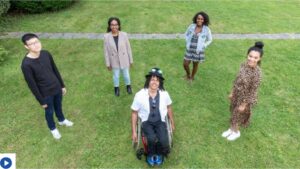 Drei Frauen und ein Mann stehen lächelnd mit etwas Abstand um einen grinsenden Mann im Rollstuhl herum. Vier der fünf jungen Menschen sind Schwarz.