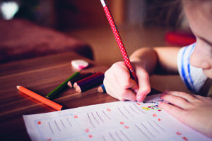 Ein Kind sitzt an einem Tisch und macht mit einem roten Bleistift Hausaufgaben.