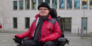 Constantin Grosch sitzt lächelnd in seinem Rollstuhl vor einem grauen Gebäude. Er trägt eine warme rote Jacke, einen Schal und einen schwarzen Hut.