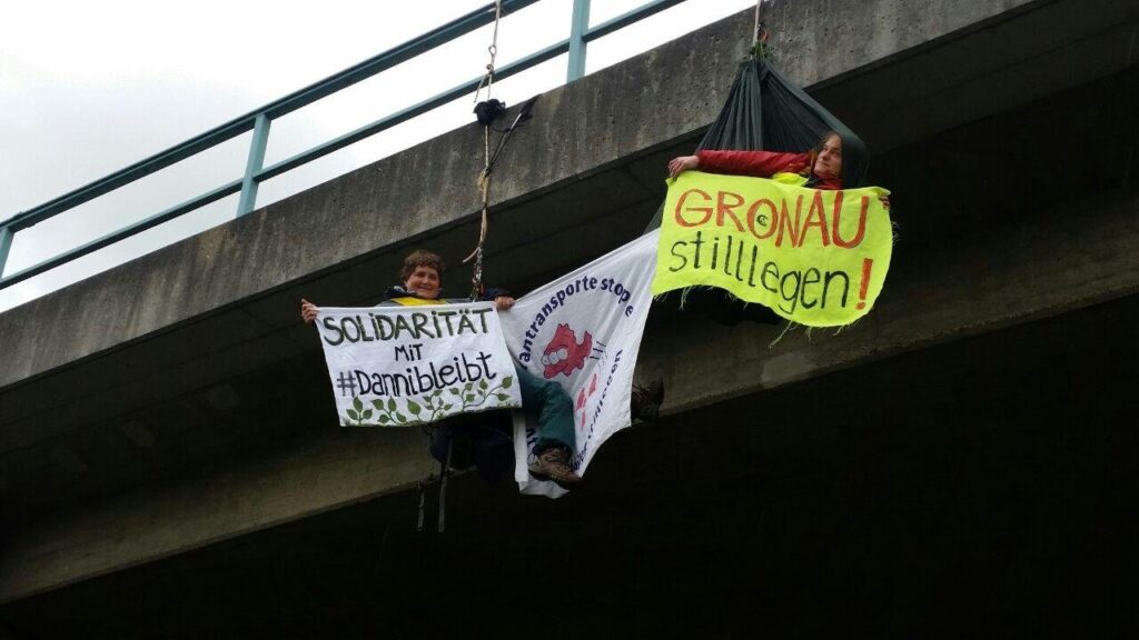 Aktivistinnen seilen sich von einer Brücke ab. Sie haben Transparente mit den Aufschriften "Solidarität mit Danni bleibt" und "Gronau stilllegen" in der Hand.