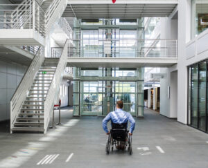 Ein weißer Mann im Rollstuhl fährt in ein großes, modernes Foyer mit vielen Treppen.