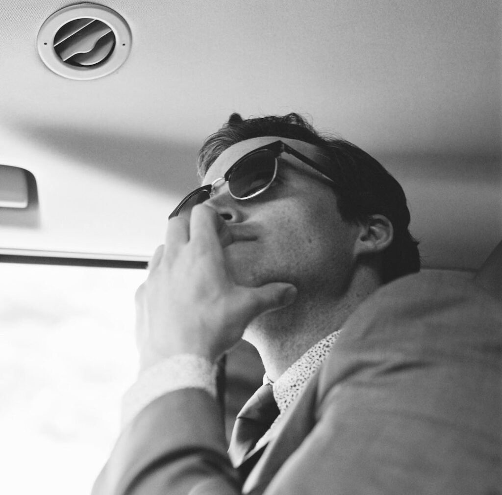 Auf einer schwarz-weiß Aufnahme sieht man einen Mann mit dunklen Haaren und Sonnenbrille aus der Froschperspektive. Er sitzt in einem Auto und schaut nachdenklich nach vorne.