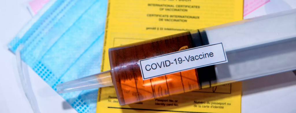 Auf einer medizinischen Maske und einem Impfausweis liegt eine gefüllte Spritze mit der Aufschrift "COVID-19-Vaccine".
