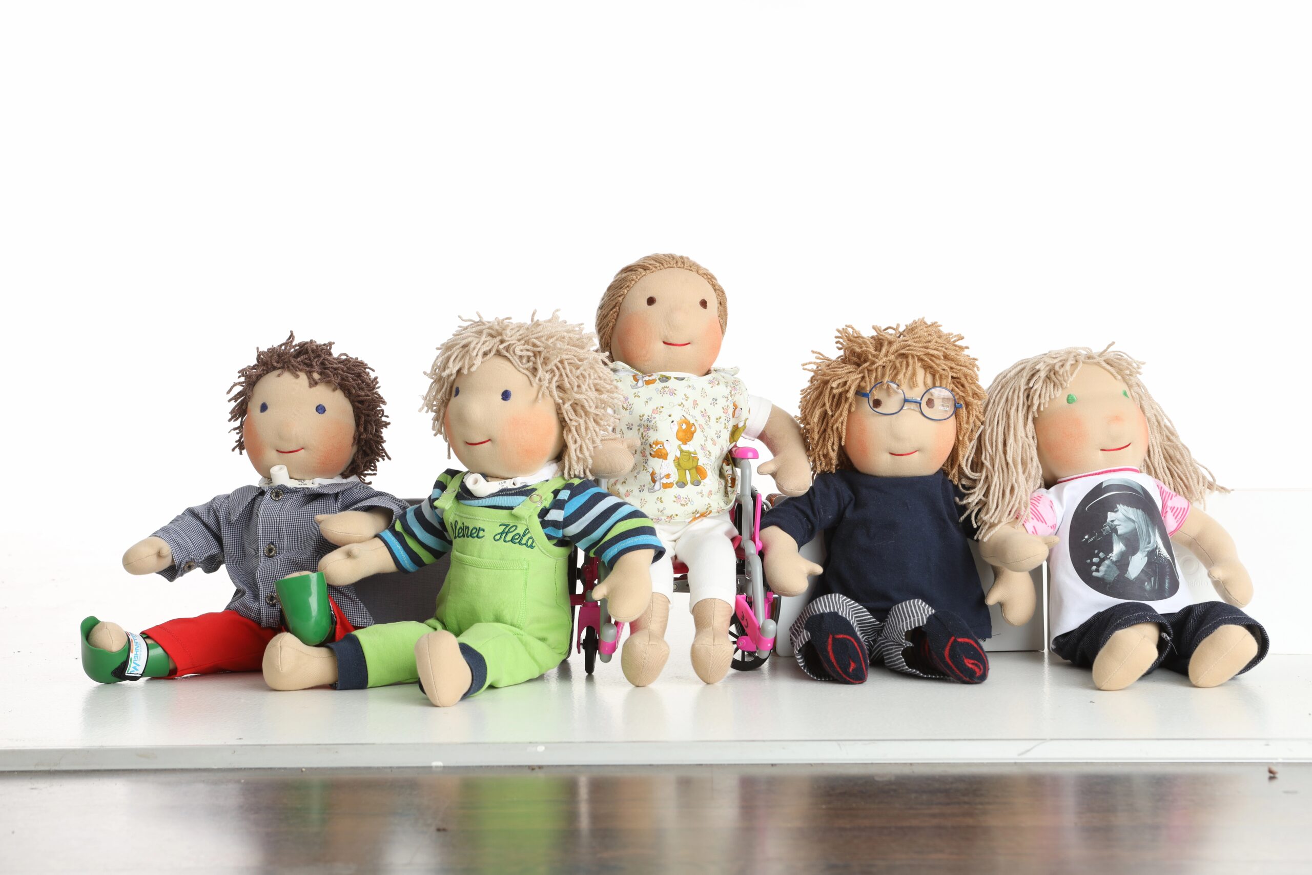 Fünf handgemachte Puppen mit Behinderungen sitzen nebeneinander. Einige tragen Orthesen, eine andere sitzt im Rollstuhl.
