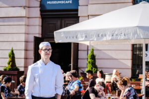 Ein junger weißer Mann in Kochkleidung steht lächelnd im Sonnenschein vor dem Museumscafe. Hinter ihm sitzen viele Gäste.