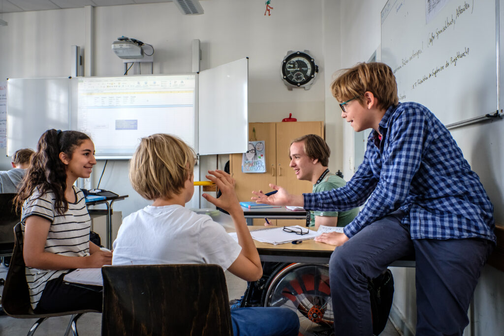 In einem Klassenraum sitzen drei Schüler:innen an einem Tisch mit einer Lehrkraft, die mit ihnen etwas bespricht.