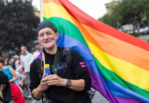 Ein älterer Mann hält auf einer Demonstration eine Regenbogenfahne in der Hand und lächelt in die Kamera.