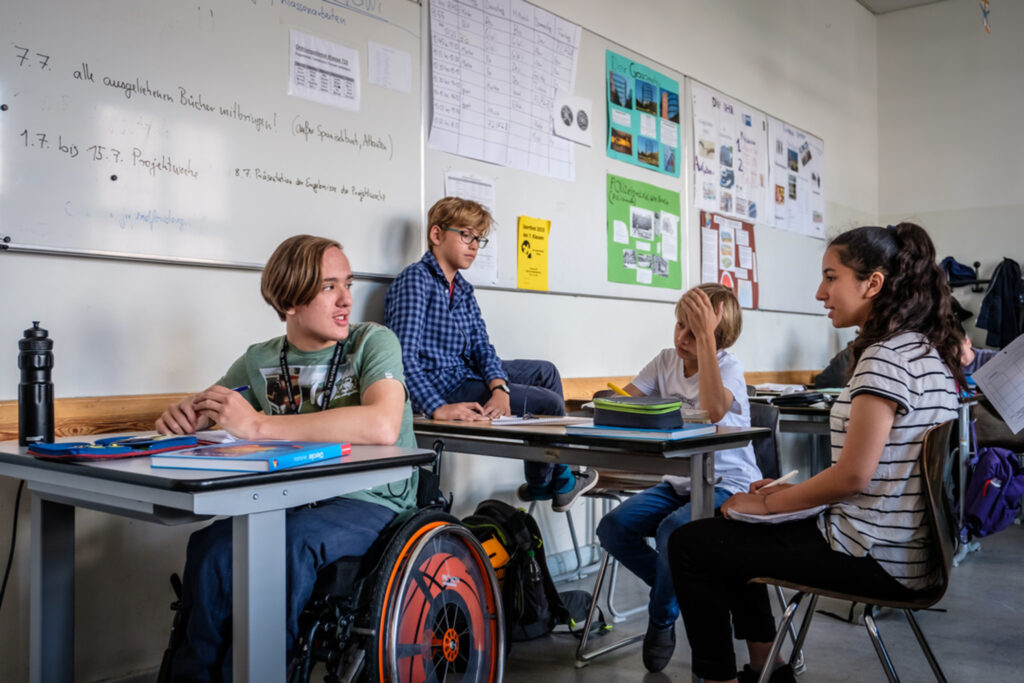 In einem Klassenzimmer reden vier Schüler+innen miteinander. Ein sSchüler sitzt im Rollstuhl.