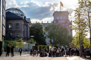 Blick auf den Reichstag in Berlin bei Sonnenschein. Davor stehen mehrere Menschen und demonstrieren.