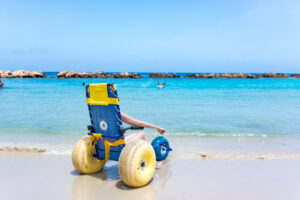 Eine Person sitzt in einem Speziellen Strand-Rollstuhl und blickt auf das kristallklare blaue Meer.