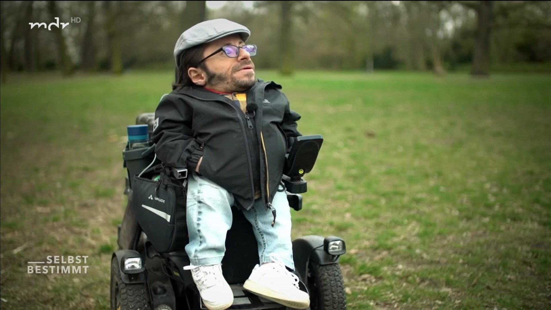 Screenshot aus der Sendung mdr Selbstbestimmt. Raul steht mit seinem Rollstuhl draussen, trägt eine graue Mütze, eine Brille und ein gelb-grau Karriertes Hemd.