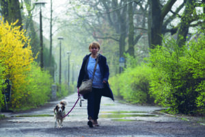 Eine Frau läuft über einen asphaltierten Waldweg auf die Kamera zu. Neben ihr läuft ein kleiner weiß-brauner Hund an der Leine.