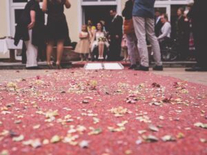 Auf einem roten Teppich liegen Rosenblätter. In der Entfernung ist ein BRautpaar, Gäste und eine Rollstuhlrampe zu sehen. Die Braut trägt ein weißes Kleid und sitzt im Rollstuhl.