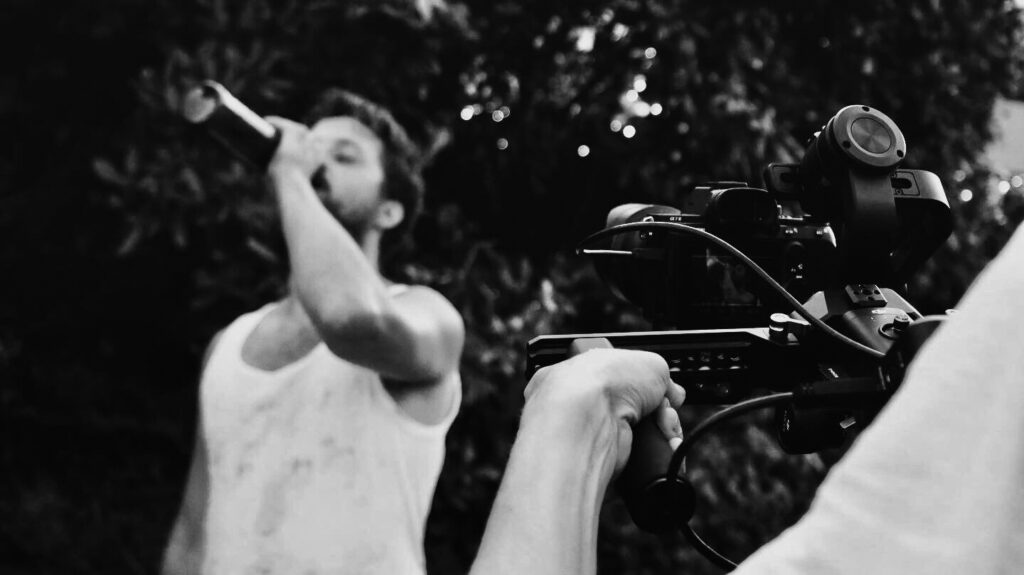 Ein Mann im weißen Unterhem wird von einer Person mit einer Kamera gefilmt. Es ist ein schwarz-weiß-Bild.