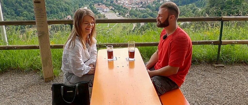 Eine kleinwüchsige Frau und ein Mann sitzen sich an einem Tisch gegenüber. Auf dem Tisch stehen getränke. Im Hintergrund der Ausblick auf ein Tal.