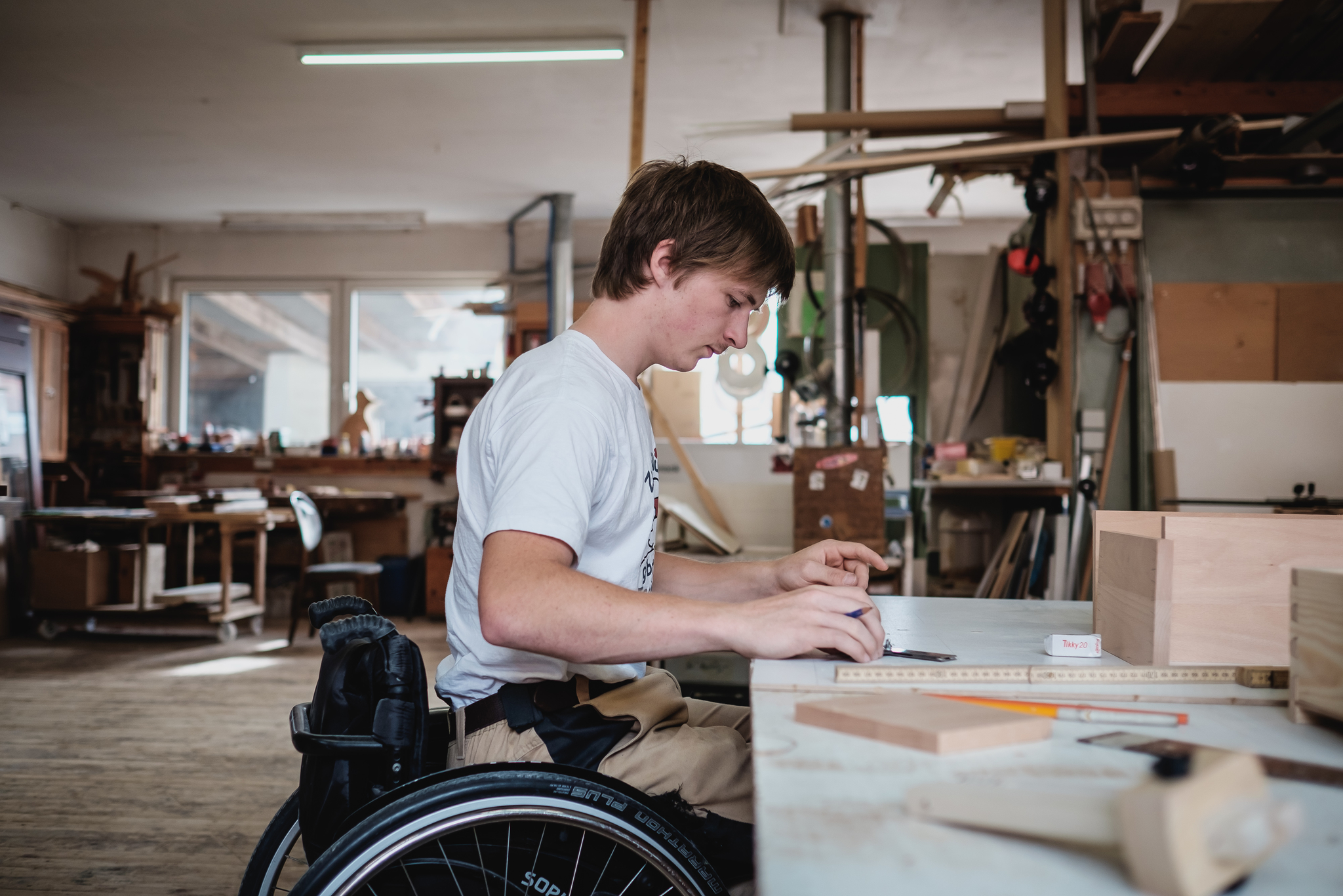 Ein junger Mann sitzt in einer Werkstatt an einem Tisch im Rollstuhl und arbeitet an Holzgegenständen.