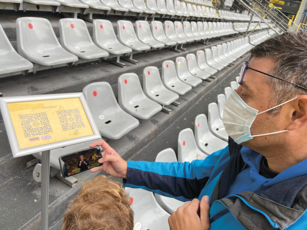 Eine Person vor Leeren Sitzbänken im Stadion bedient eine App am Smartphone.