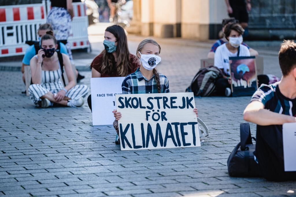 Greta Thunberg sitzt auf einem Platz und hat ihr Schiild mit der Aufschrift "Schulstreik für das Klima" vor sich. Um sie herum sitzen andere Jugendliche die ebenfalls schilder halten. Alle Tragen einen Mund-Nase-Schutz.