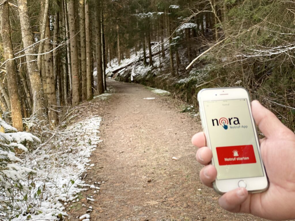 Auf einem Waldweg hält eine Hand ein Smartphone, auf dem die nora-App geöffnet ist.