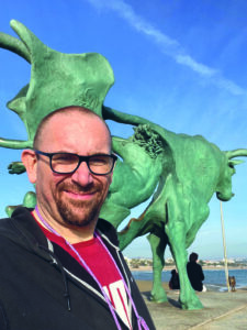 Ein Mann mit Halbglatze, Brille und Bart steht vor einer grünen Statue und schaut in die Kamera.