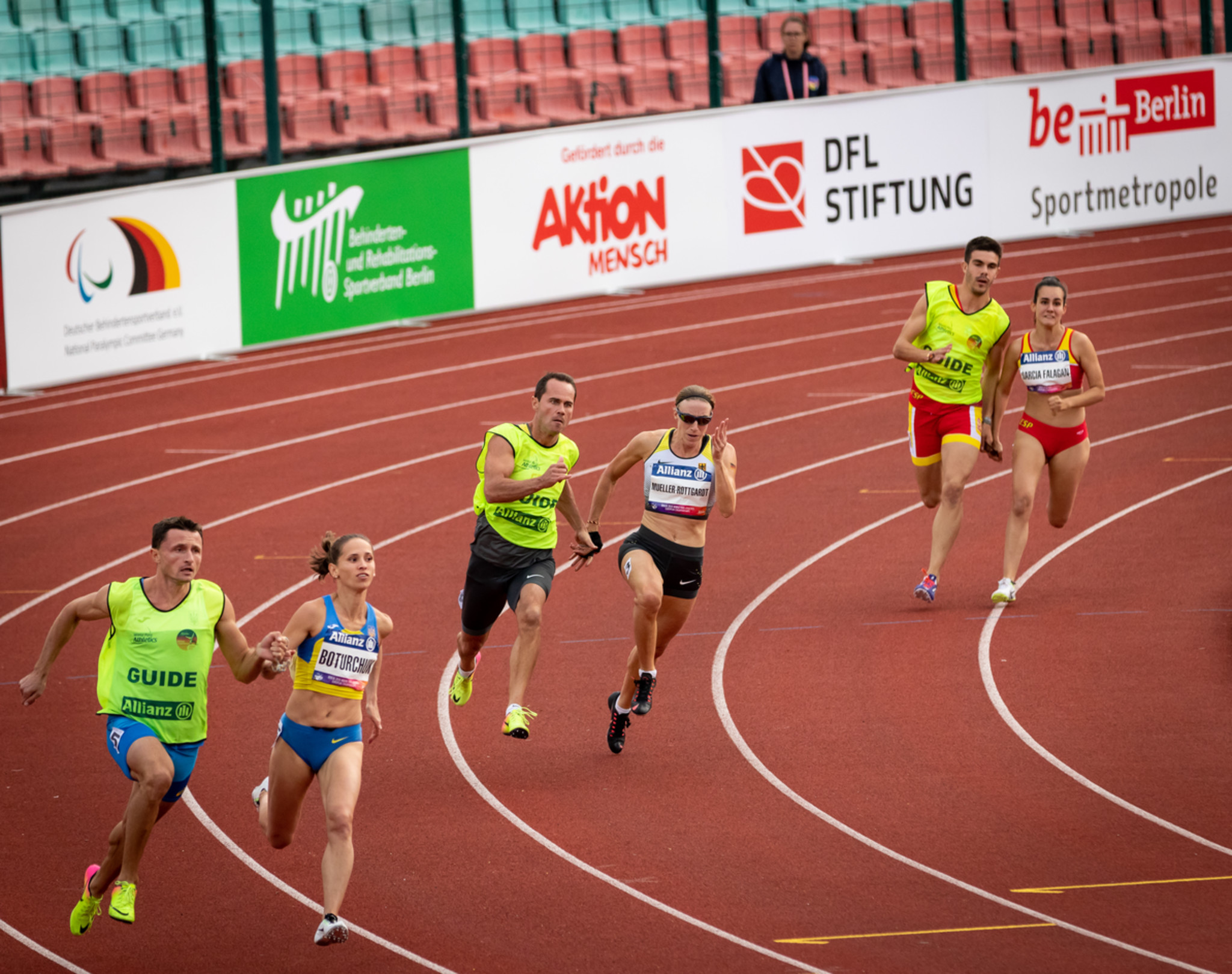 Auf einer Laufbahn biegen drei blindeLäuferinnen mit ihren Assistenz-Läufern auf die Zielgerade ein.