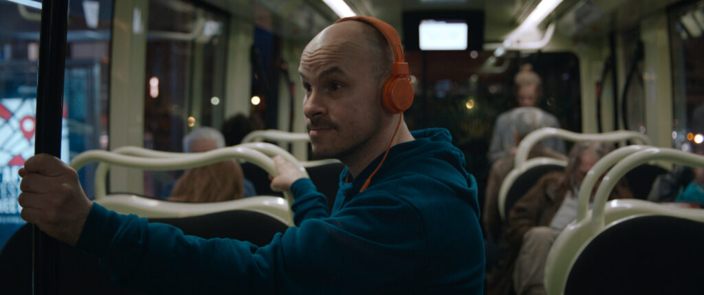 Igor, ein weißer Mann mit Glatze steht im Bus und hat orange Kopfhörer auf.