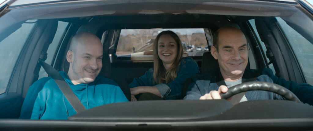 Im Innenraum eines Autos sitzen zwei lachende Männer und hinten eine grinsende Frau.