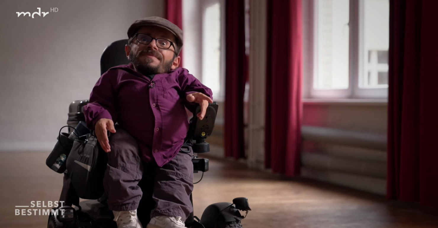 Foto von Raul Krauthausen. Er sitzt im Rollstuhl, trägt ein violettes Hemd und schaut in die Kamera.
