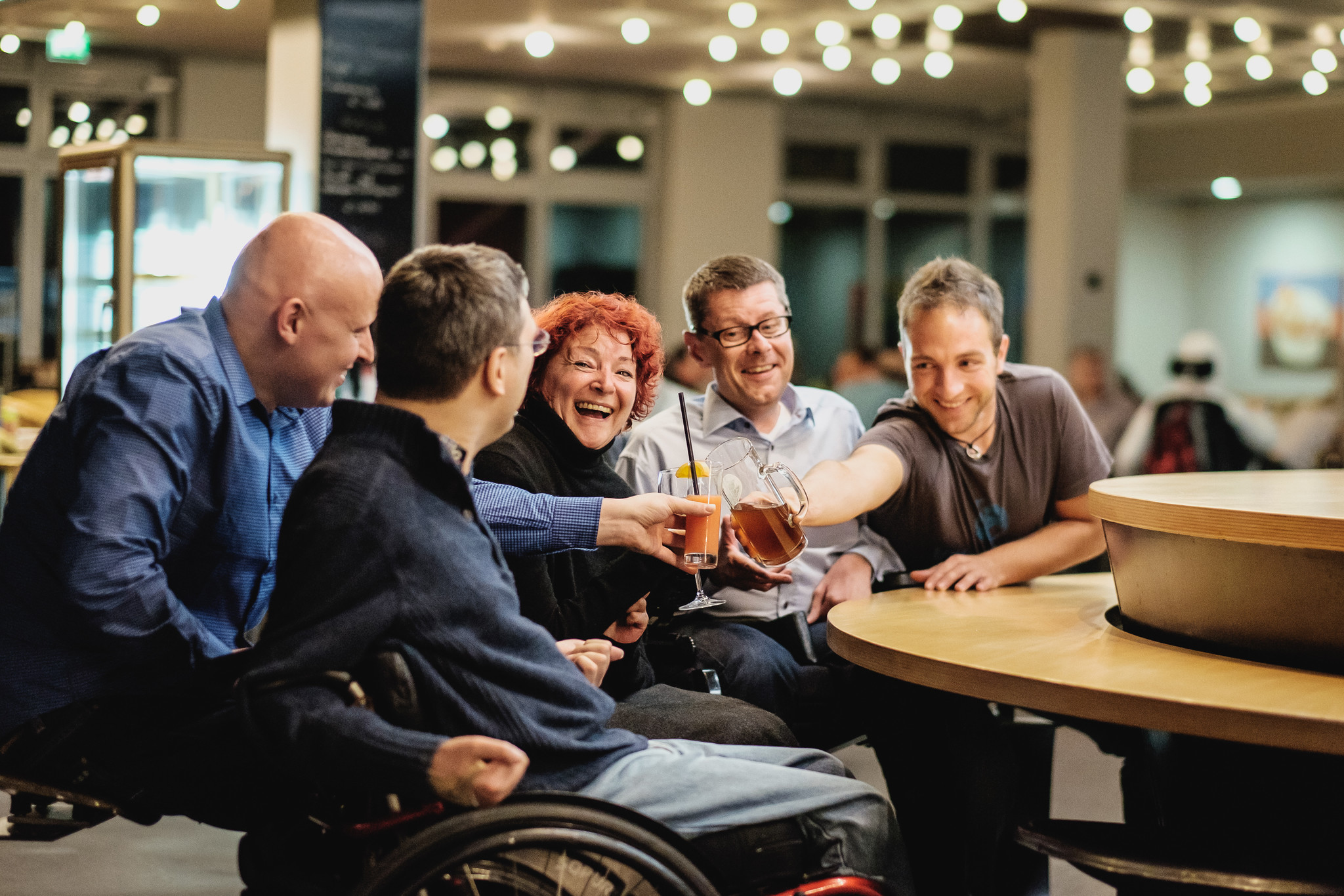 Mehrere Menschen mit Behinderung sitzen zusammen, stoßen mit Getränken an und lachen fröhlich.