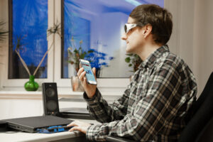 Ein blinder Mann trägt eine Sonnenbrille, sitzt in einem Büro am Schreibtisch und hält ein Smartphone in der Hand.