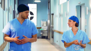 Kassandra Wedel steht neben einem Schauspielkollegen, im Krankenhaus. Beide schauen sich an. Sie tragen beide blaue OP Klamotten.