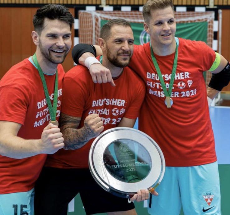 Federico mit zwi Mitspielern. Sie halten jubelnd die Meisterschale in ihren Händen, tragen rote T-Shirts mit der Aufschrift "Deutscher Futsal Meister 2021 und haben eine Medallie um den Hals hängen.