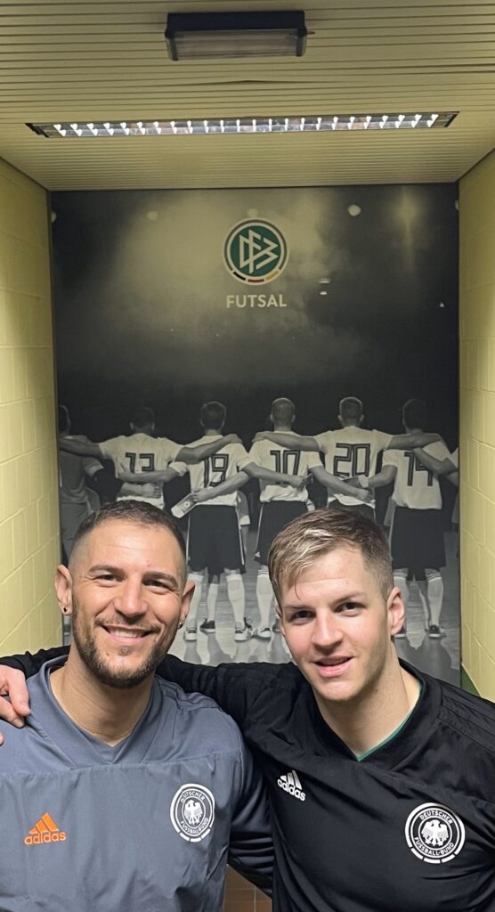 Stefano und Philipp sitzen Arm in Arm vor einem Poster des DFB und schauen in die Kamera.