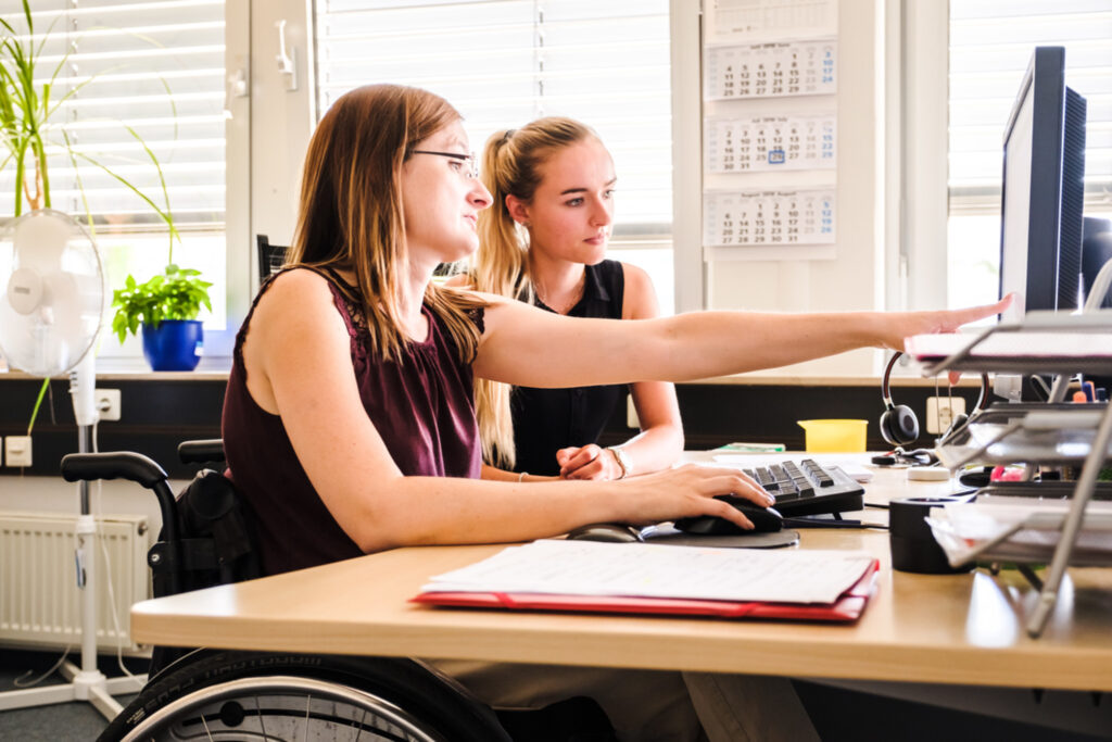 Eine Frau sitzt im Rollstuhl und arbeitet an Computer. Sie zeigt ihrer Kollegin ohne Behinderung, die neben ihr sitzt, etwas auf dem Bildschirm.