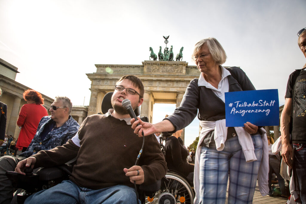 Eine Person sitzt im Rollstuhl vor dem Brandenburger Tor in Berlin und hält eine Rede. Ihr wird von einer anderen Person das Mikro gehalten.