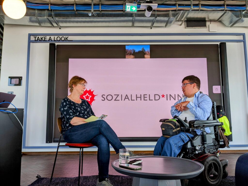 Eine Frau ohne Behinderung interviewt einen Mann, der in einem Elektrorollstuhl sitzt.
