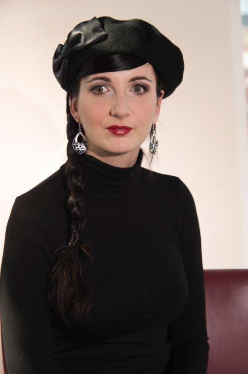 Portrait von Jennifer Sonntag, einer weißen Frau mit schwarzen Haaren, die in einem seitlichen Flechtzopf über ihre Schulter nach vorne hängen. Sie trägt einen schwarzen Rollkragenpullover und und einen schimmernden, schwarzen Hut mit seitlicher Schleife.