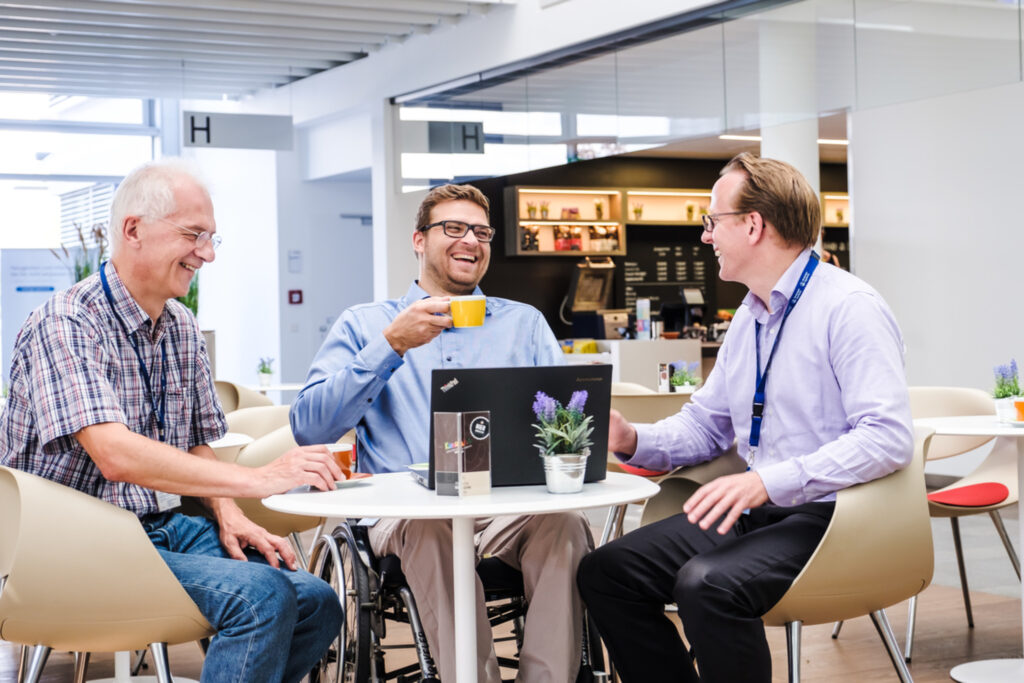 Drei Männer sitzen an einem Tisch, auf dem ein Laptop steht. Eine Person sitzt im Rollstuhl, eine trinkt etwas aus einer Tasse, alle lachen.